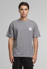 Gray cotton t-shirt with a loose fit, short sleeves, and a white logo on the left chest. Smooth texture, simple design, no patterns.