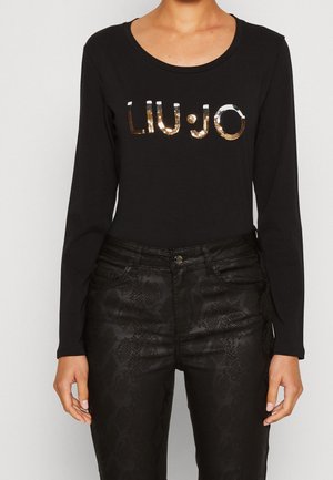 Femme portant un haut noir à manches longues avec un logo "LIU·JO" en sequins et un pantalon noir taille haute avec un motif serpent subtil.