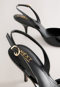 Sandales à talons hauts en cuir verni noir avec fines lanières et boucles dorées sur fond neutre.