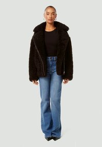 Casaco de pêlo sintético preto com gola larga e fecho éclair, usado sobre uma blusa preta e jeans azuis à boca de sino, colocado contra um fundo neutro.