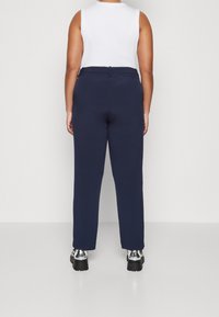 Personne portant un pantalon bleu marine, un haut sans manches blanc et des chaussures à plateforme argentées brillantes, se tenant de dos contre un fond uni.