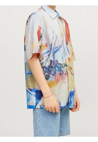 Henkilö, joka on pukeutunut löysään lyhythihaiseen napitettavaan paitaan, jossa on abstrakti monivärinen kuosi, ja vaaleansinisiin farkkushortseihin, seisoo kädet rentoina.