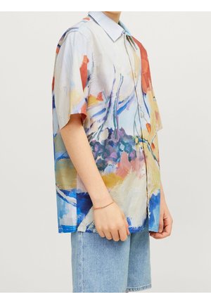 Personne portant une chemise à manches courtes ample et à boutons avec un motif abstrait multicolore et un short en denim bleu clair, se tenant avec les mains détendues.