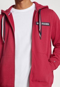 Haut zippé rouge en coton doux, avec un patch logo contrastant, des cordons de tirage bicolores et des poches avant. T-shirt blanc visible en dessous.