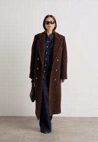 Brūns vilnas mētelis ar divrindu dizainu, lieliem apkakles galiem un sānu kabatām, uzvilkts virs tumšas kreklu un džinsiem. Komplektā ietilpst saulesbrilles un brūns soma.