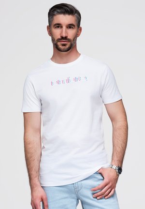 Homme aux cheveux courts et foncés et barbe porte un t-shirt blanc avec un texte coloré et un jean bleu clair, debout devant un fond uni.