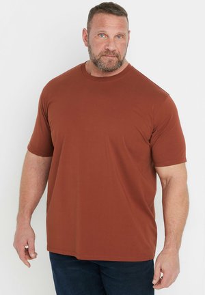 Voksen mand med kort hår og skæg iført en ensfarvet rustfarvet kortærmet skjorte og mørke bukser, stående mod en hvid baggrund.