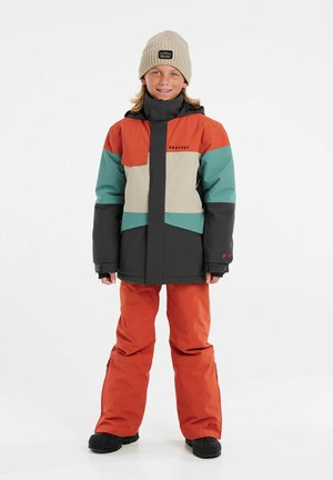 Abrig colorido de invierno en naranja, verde azulado y gris, con un panel central beige. Combinado con pantalones naranjas y un gorro de punto beige.