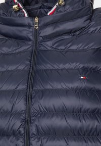 Marineblaues Steppjacke mit Reißverschluss vorne, ausgestattet mit einem gecoachten Muster, goldenen Akzenten und einem kleinen Logotag in Rot, Weiß und Blau auf der unteren linken Seite.