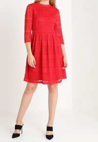 Robe en dentelle rouge avec des manches trois-quarts, des motifs texturés et une jupe évasée. Associée à des chaussures à talons noires et à un décolleté festonné.