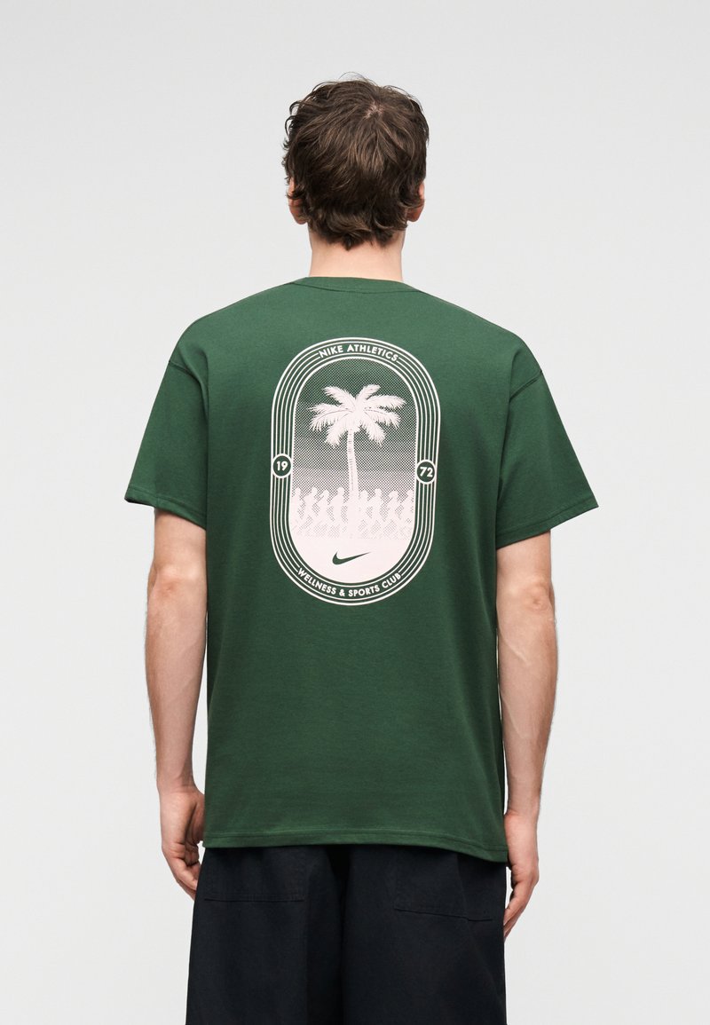 Άνδρας που φοράει σκούρο πράσινο T-shirt με λευκό σχέδιο φοίνικα και λογότυπο Nike Athletics στην πλάτη, συνδυασμένο με μαύρο σορτς, στέκεται με την πλάτη γυρισμένη.