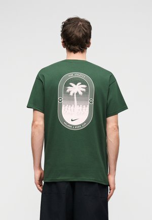 Man draagt een donkergroen T-shirt met een witte palmboom en Nike Athletics-logo op de achterkant, gecombineerd met een zwarte short, en staat met de rug naar de camera.