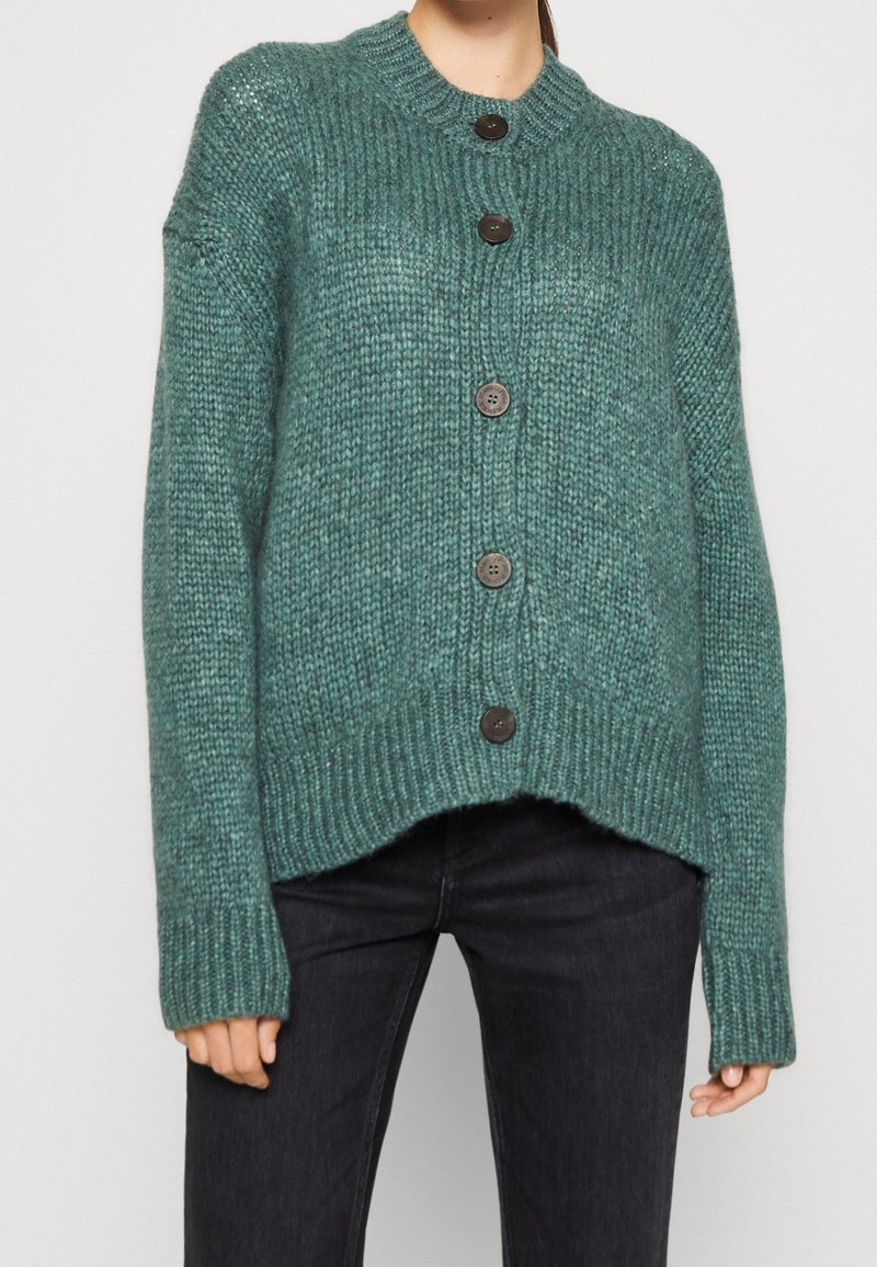 Gilet en tricot turquoise avec un col rond, cinq boutons foncés, texture côtelée et coupe décontractée. L'ourlet court ajoute de la dimension.