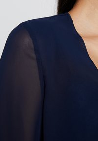 Blusa navy con scollatura a V, maniche lunghe trasparenti e tessuto testurizzato. Rifinita con una superficie liscia lungo il colletto.