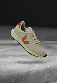 Joggesko med en beige og kremet mesh-overdel, oransje aksenter, og en teksturert gummisåle med minimalistisk design og synlige lisser.