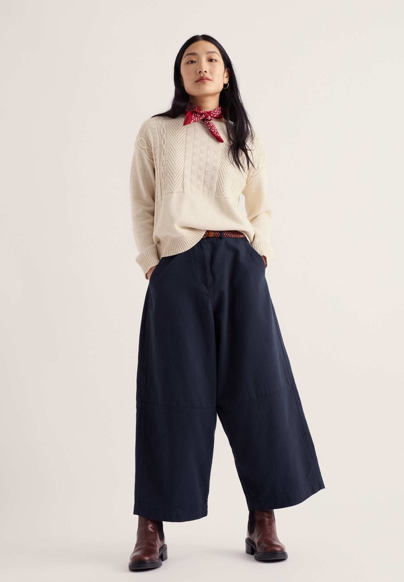 Femme portant un pull en maille crème, un foulard rouge autour du cou, un pantalon large bleu marine, des bottines marron, debout les mains dans les poches devant un fond uni.