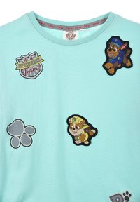 Helles, blaues Kurzarmhemd mit verschiedenen Aufnähern: einem Wappen-Logo, einem Pfotenabdruck und cartoonhaften Hundekaraktern in verspielten Posien.