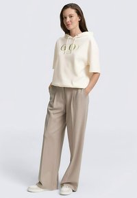 Kremowa bluza z kapturem z logo, krótkie rękawy, w zestawieniu z beżowymi szerokimi spodniami i białymi trampkami. Gładka tkanina, luźny krój.