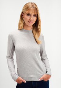 Kobieta z blond włosami ubrana w jasnoszary sweter z długim rękawem i ciemnoniebieskie dżinsy, z rękami w kieszeniach, delikatnie uśmiechająca się na tle białego tła.