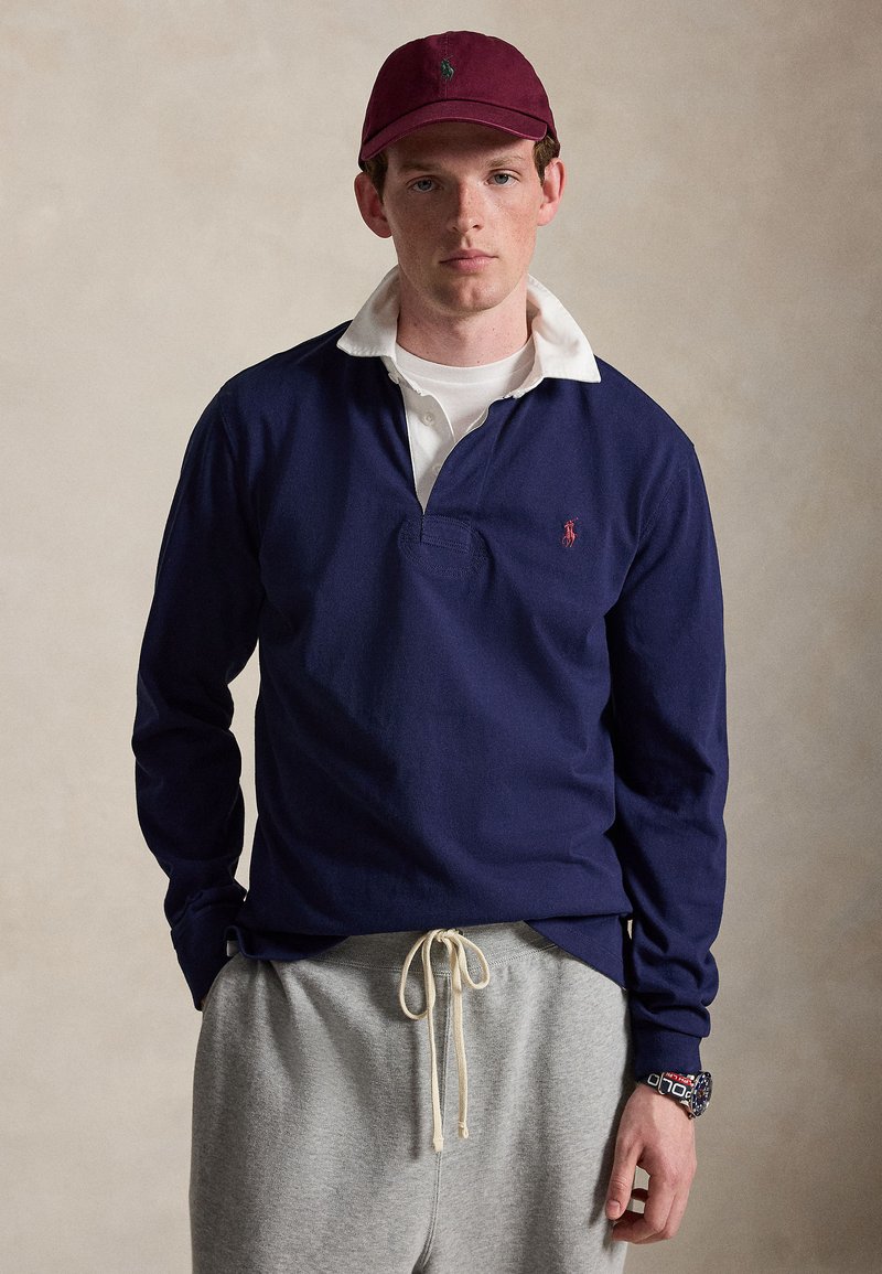 Ung mand iført en mørkerød kasket, navyblå langærmet rugbyshirt med hvid krave, grå joggingbukser og et stort sort armbåndsur, stående.