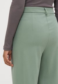 Helle grüne weit geschnittene Hose aus glattem Material, mit einem vorderen Knopfverschluss und einem taillierten Bund, kombiniert mit einem figurbetonten dunkelgrauen Oberteil.
