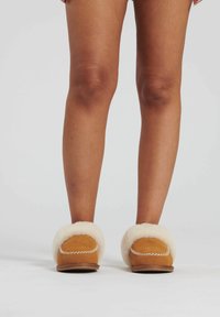Brune ruskinds slippers med hvidt pelsfor, der har en syet kant og flad sål. Billedet viser slippers iført, med fokus på benene og fodtøjet.