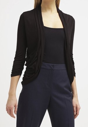 Black draped cardigan with long sleeves, made from soft fabric. Paired with fitted navy trousers, showcasing a simple, elegant silhouette.