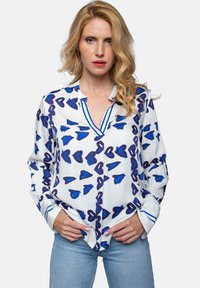 Biała bluzka w niebieskie serca, z dekoltem w serek, długimi rękawami i kontrastującymi niebieskimi paskami wzdłuż kołnierza i mankietów.