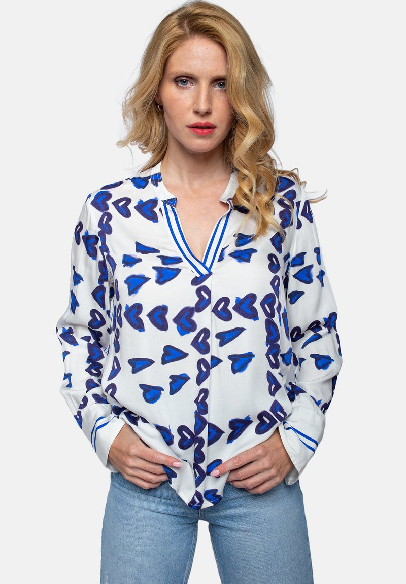 Biała bluzka w niebieskie serca, z dekoltem w serek, długimi rękawami i kontrastującymi niebieskimi paskami wzdłuż kołnierza i mankietów.