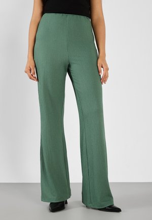 Groene, gestructureerde wijdpantalon met een hoge taille en zijsplitten, gecombineerd met zwarte puntige schoenen tegen een neutrale achtergrond.