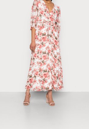 Nainen valkoisessa kietaisumekossa, jossa on vaaleanpunainen kukkakuosi ja kolme neljäsosan mittaiset hihat, yhdistettynä vaaleanpunaisiin korollisiin sandaaleihin.