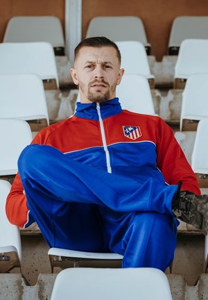 Man zit met gekruiste benen op lege stoelen in het stadion, draagt een rood-blauw trainingspak met een sportteamlogo.
