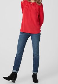 Rode blouse met lange mouwen en een keyhole-halsdetail, gecombineerd met blauwe jeans met opgerolde mouwen en zwarte puntige enkellaarsjes.