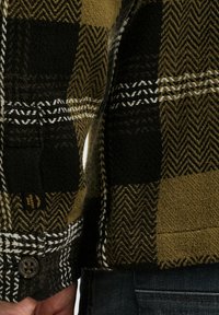 Geknoopte flanellen shirt in zwart, olijf en crèmekleurig ruitpatroon. Kenmerkt zich door een gestructureerde stof, een onderscheidend visgraatontwerp en een merketiket.