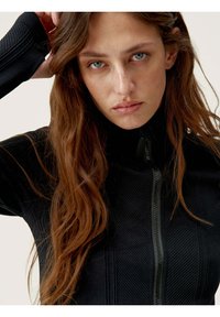 Black zip-up jacket made of textured fabric with vertical stripes. High collar and fitted sleeves. Model has long brown hair.