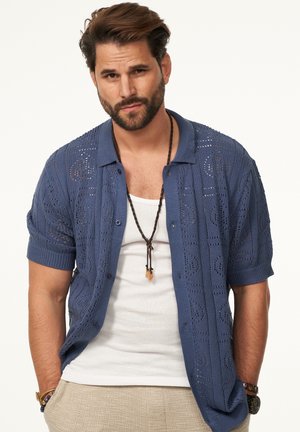 Blauses, kurzärmeliges Strickhemd mit strukturiertem, durchbrochenem Muster, Knopfverschluss und Kragen, getragen über einem weißen Tanktop.