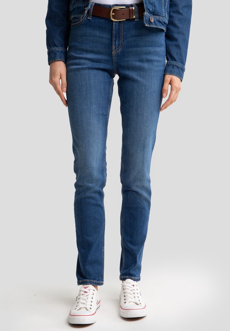 Personne portant un jean en denim bleu, une ceinture marron, une veste en denim et des baskets blanches, debout contre un fond uni.