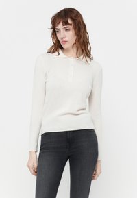 Lys beige langermet genser med polo-krage, med en knappestolpe og en myk, strikket tekstur. Kombinert med mørkegrå jeans.