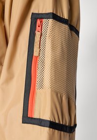 Beige jakke med svart trim har en mesh-lomme og en kontrasterende oransje glidelås på siden, som viser varierte teksturer og materialer.