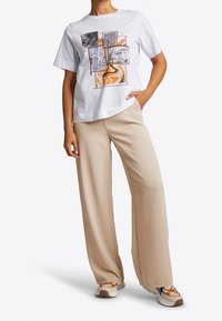 Camiseta blanca de algodón con diseños gráficos multicolores, combinada con pantalones de pierna ancha beige y zapatillas con patrón.