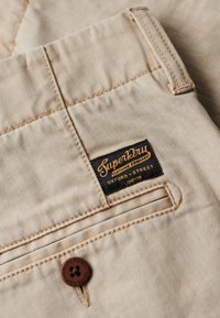 Tessuto di denim beige con cuciture visibili, dotato di un'etichetta nera con testo dorato. Include un bottone marrone con il nome del marchio.