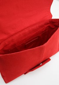 Punainen kangaspussi, jossa on taitettavan läpän muoto, sisätasku ja sileä pinta. Sulkeutuu nepparilla.