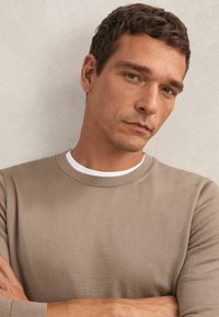 Beżowa koszula z długim rękawem o fakturowanym wykończeniu, z okrągłym dekoltem, noszona na białej koszulce. Model pozujący ze skrzyżowanymi ramionami na neutralnym tle.