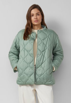 Kobieta w jasnozielonej, pikowanej kurtce na kremowym swetrze i białych spodniach, stojąca z rękami w kieszeniach na szarym tle.