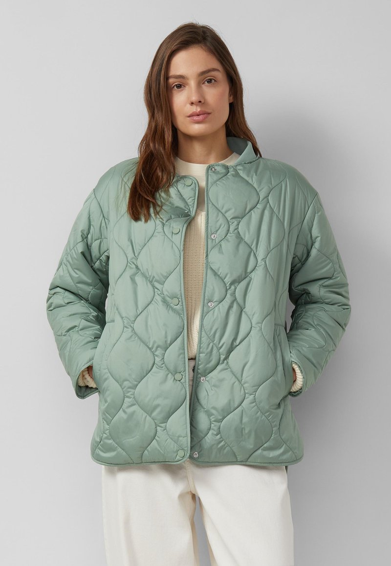 Femme portant une veste matelassée vert clair par-dessus un pull crème et un pantalon blanc, debout les mains dans les poches devant un arrière-plan gris.