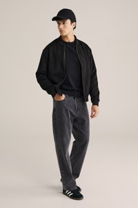 Melnas zamšādas bomber jakas, melna džemperis, pelēki džinsi un melni sporta apavi ar baltām svītrām. Modelis stāv pret neitrālu fonu.