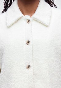 Gros plan d'une veste blanche texturée à boutons avec un col, montrant trois gros boutons ronds et une partie du cou et des cheveux d'une personne.