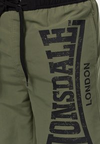 Olīvu zaļi šorti ar melnu elastīgu jostu, kuriem ir liels nolietots melns druku logo sānos. Teksturēts, viegls audums.