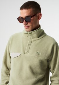 Hellgrüner Fleece-Pullover mit hohem Kragen, Knopfverschluss und einer cremefarbenen aufgesetzten Tasche. Bestickte Kreisdetaillierungen auf der Vorderseite.