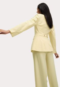 Yellow tailored suit with long sleeves and a peplum waist. Features two buttons at the back and wide-leg trousers. Smooth fabric texture.